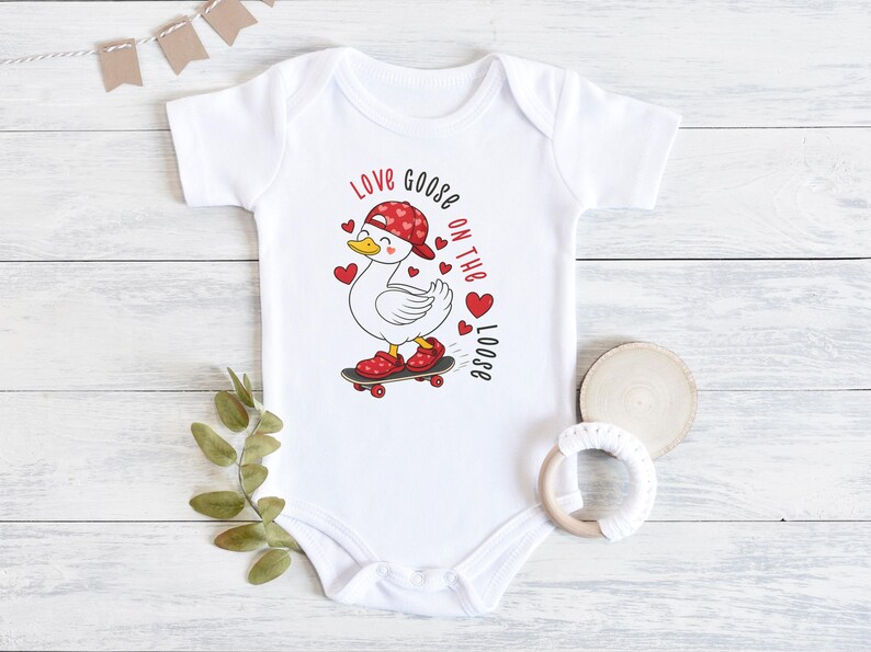 May include: White baby onesie with a cartoon goose wearing a red hat and shoes, riding a skateboard. The text "Love Goose on the Loose" is printed in red. The onesie has short sleeves and a round neckline.