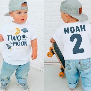 May include: A toddler wearing a white t-shirt with the text "TWO the MOON" and space-themed graphics. The child is also wearing light blue jeans and a gray baseball cap. The back of the shirt has the name "NOAH" and the number "2".