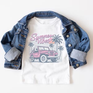 Puede incluir: Camiseta blanca con un gr&aacute;fico de un jeep rosa y palmeras. El texto "Summer Vibes" est&aacute; impreso en rosa en la camiseta.