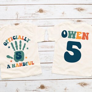 May include: Two white t-shirts with a retro design. The front shirt says "Officially a Handful" with a handprint and the number 5. The back shirt says "Owen" with the number 5.