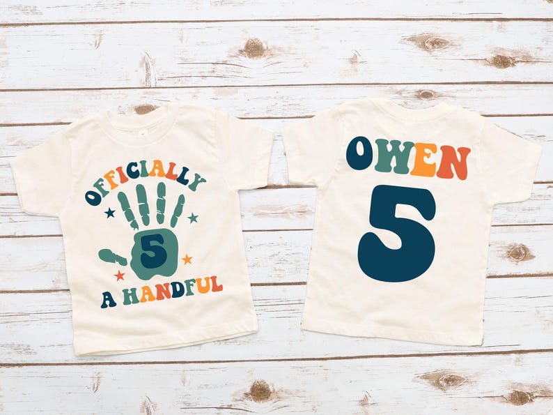 May include: Two white t-shirts with a retro design. The front shirt says "Officially a Handful" with a handprint and the number 5. The back shirt says "Owen" with the number 5.