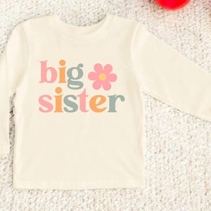 Puede incluir: Camiseta de manga larga color crema con las palabras "big sister" en letras coloridas de estilo retro. Un gr&aacute;fico de flor rosa est&aacute; incorporado en el dise&ntilde;o. La camiseta est&aacute; hecha de un material suave.