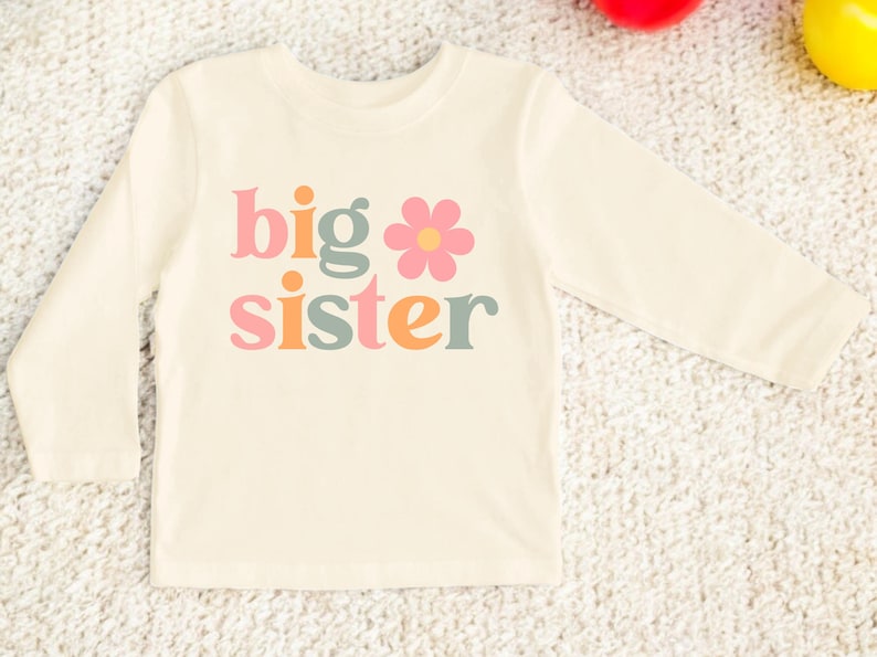 Puede incluir: Camiseta de manga larga color crema con las palabras "big sister" en letras coloridas de estilo retro. Un gr&aacute;fico de flor rosa est&aacute; incorporado en el dise&ntilde;o. La camiseta est&aacute; hecha de un material suave.