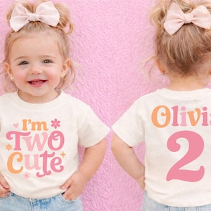 May include: A toddler wearing a cream-colored t-shirt with the text "I'm Two Cute" in pink, orange, and cream. The back of the shirt says "Olivia 2" in matching colors. The child has a pink bow in their hair.