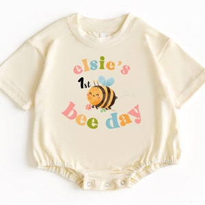 May include: A cream-colored baby onesie with a bee graphic and the text "elsie's 1st bee day" in colorful letters.