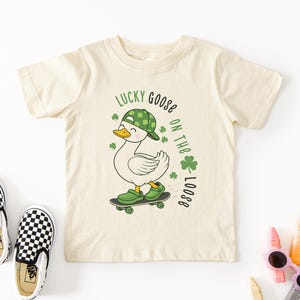 May include: Cream-colored toddler t-shirt with a graphic of a skateboarding goose wearing a green hat and shoes. The text "Lucky Goose on the Loose" is printed in green. Black and white checkered slip-on shoes are next to the shirt.
