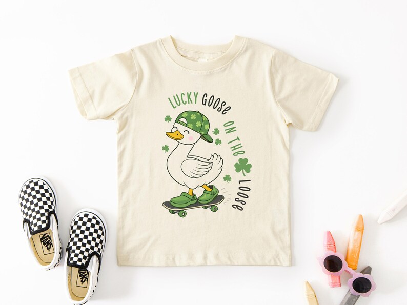 May include: Cream-colored toddler t-shirt with a graphic of a skateboarding goose wearing a green hat and shoes. The text "Lucky Goose on the Loose" is printed in green. Black and white checkered slip-on shoes are next to the shirt.