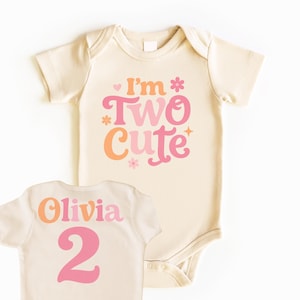 May include: Cream-colored baby bodysuit with the text "I'm Two Cute" in pink and orange. The back of the bodysuit has the name "Olivia" and the number "2" in pink. A cute outfit for a second birthday.