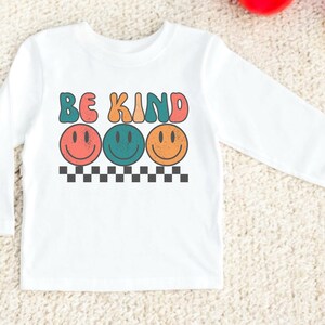 May include: White long-sleeve shirt with the text "Be Kind" in colorful letters and three smiling faces in different colors.