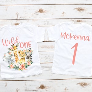 May include: Two white t-shirts with a pink design. The front of one shirt says "Wild One" with a giraffe, lion, zebra, and elephant. The back of the other shirt says "Mckenna 1".