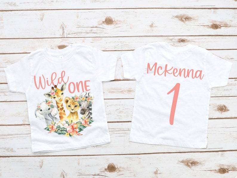 May include: Two white t-shirts with a pink design. The front of one shirt says "Wild One" with a giraffe, lion, zebra, and elephant. The back of the other shirt says "Mckenna 1".