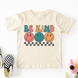 May include: A beige short-sleeved t-shirt with the text "BE KIND" in colorful retro letters and three colorful smiley faces below the text. The shirt is on a white background with black and white checkered Vans shoes and colorful crayons.