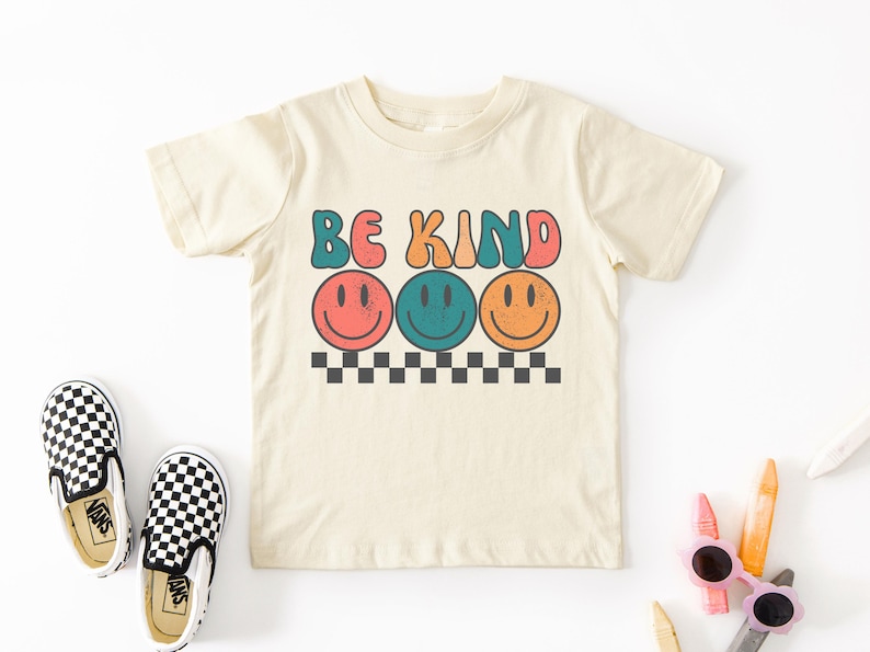 May include: A beige short-sleeved t-shirt with the text "BE KIND" in colorful retro letters and three colorful smiley faces below the text. The shirt is on a white background with black and white checkered Vans shoes and colorful crayons.
