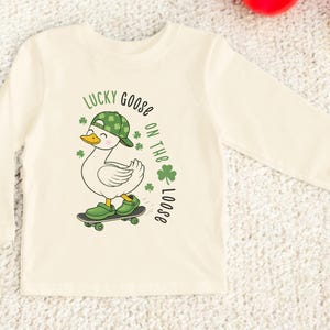 May include: Cream-colored long-sleeved shirt featuring a cartoon goose on a skateboard. The goose wears a green hat with shamrocks and green shoes. Text reads "Lucky Goose on the Loose" with shamrock accents.