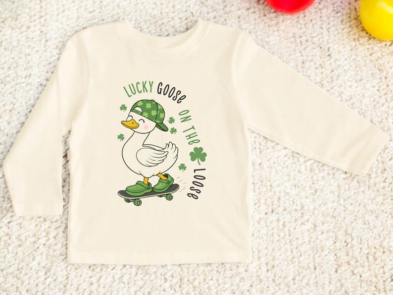 May include: Cream-colored long-sleeved shirt featuring a cartoon goose on a skateboard. The goose wears a green hat with shamrocks and green shoes. Text reads "Lucky Goose on the Loose" with shamrock accents.
