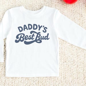 May include: White long-sleeve shirt with the text "DADDY'S Best Bud" in navy blue. The shirt is made of a soft material and is suitable for children. The design is simple and the shirt is likely for casual wear.