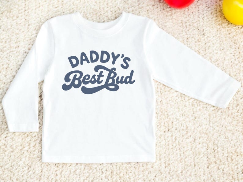 May include: White long-sleeve shirt with the text "DADDY'S Best Bud" in navy blue. The shirt is made of a soft material and is suitable for children. The design is simple and the shirt is likely for casual wear.