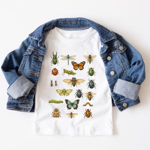 May include: A white t-shirt featuring a colorful illustration of various insects, including butterflies, beetles, and a dragonfly. The shirt is partially covered by a blue denim jacket.