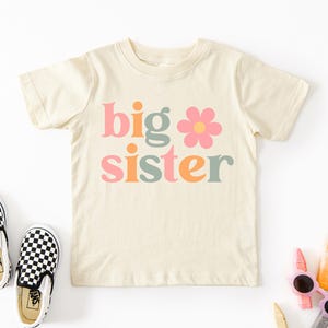 Puede incluir: Camiseta crema para ni&ntilde;os peque&ntilde;os con las palabras "big sister" en rosa, naranja y verde, con una flor. Zapatos sin cordones a cuadros blancos y negros a la izquierda. Crayones y gafas de sol a la derecha.