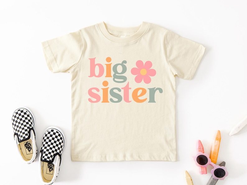 Puede incluir: Camiseta crema para ni&ntilde;os peque&ntilde;os con las palabras "big sister" en rosa, naranja y verde, con una flor. Zapatos sin cordones a cuadros blancos y negros a la izquierda. Crayones y gafas de sol a la derecha.