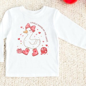 May include: White long-sleeved shirt featuring a cartoon goose wearing pink boots and a bow. The goose is surrounded by hearts and gift boxes. Text on the shirt reads "Lovely Goose on the Loose".