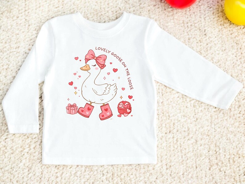 May include: White long-sleeved shirt featuring a cartoon goose wearing pink boots and a bow. The goose is surrounded by hearts and gift boxes. Text on the shirt reads "Lovely Goose on the Loose".