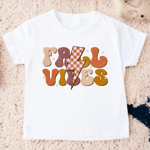 May include: White t-shirt with a retro-style graphic that says "Fall Vibes" in orange, brown, and pink letters with a checkered lightning bolt in the middle.