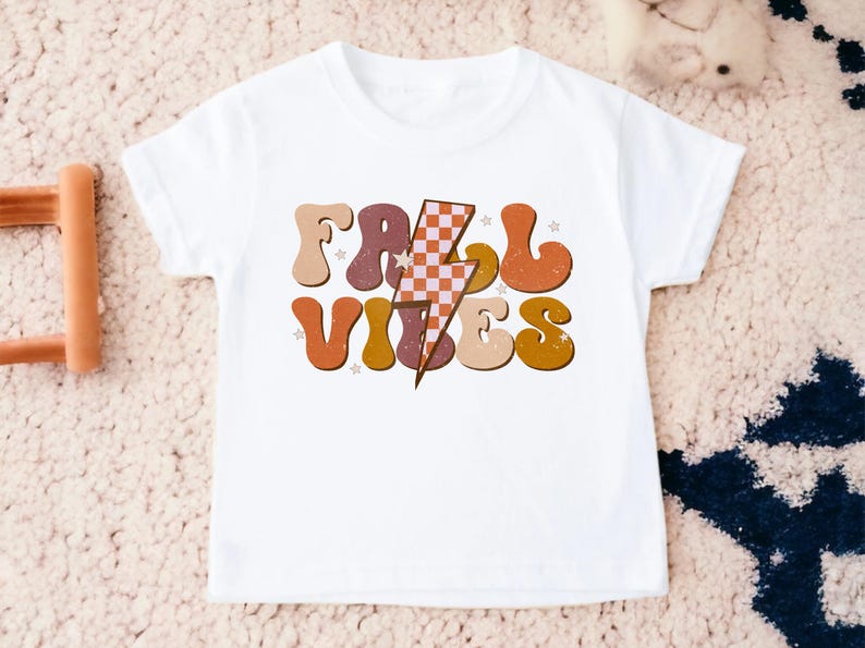 May include: White t-shirt with a retro-style graphic that says "Fall Vibes" in orange, brown, and pink letters with a checkered lightning bolt in the middle.