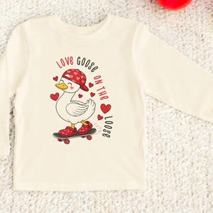 May include: Cream-colored long-sleeved shirt featuring a cartoon goose on a skateboard, wearing a red cap and shoes with heart patterns. The text "Love goose on the loose" is printed in red.