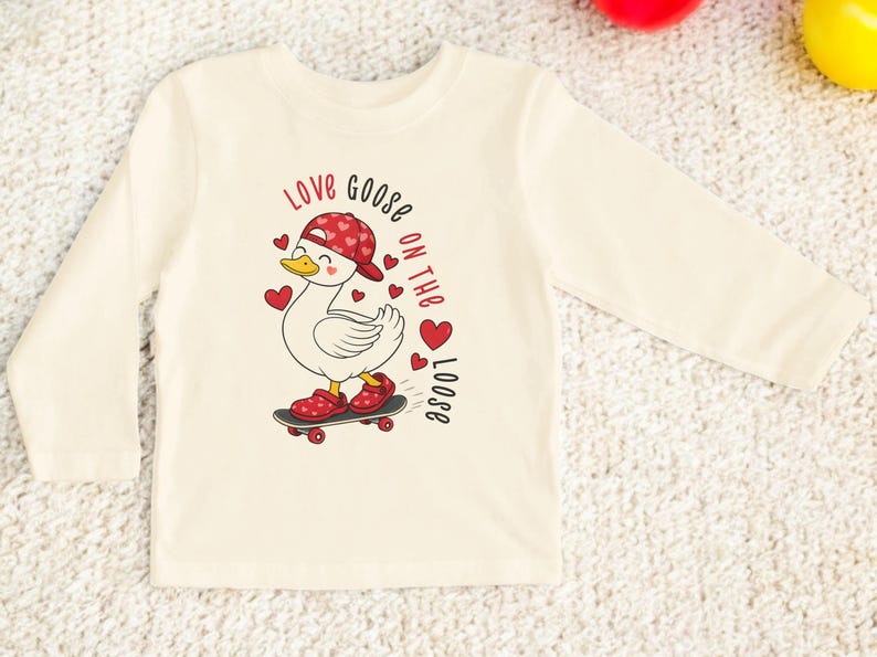 May include: Cream-colored long-sleeved shirt featuring a cartoon goose on a skateboard, wearing a red cap and shoes with heart patterns. The text "Love goose on the loose" is printed in red.