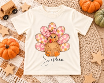 May include: A cream-coloured short-sleeve t-shirt featuring a central graphic of a brown turkey with a pink bow and tail feathers patterned with pink and orange checkered and floral designs, with the name Sophia written in black script below.