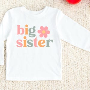 Puede incluir: Camiseta blanca de manga larga con las palabras "big sister" en letras rosas, verdes y naranjas, y un gr&aacute;fico de una flor rosa. La camiseta est&aacute; hecha de un material suave y es adecuada para ni&ntilde;os.