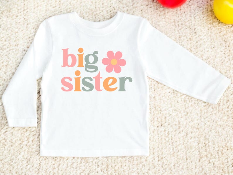 Puede incluir: Camiseta blanca de manga larga con las palabras "big sister" en letras rosas, verdes y naranjas, y un gr&aacute;fico de una flor rosa. La camiseta est&aacute; hecha de un material suave y es adecuada para ni&ntilde;os.