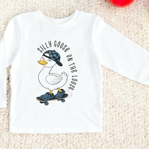 May include: White long-sleeved top with a cartoon duck wearing a cap and shoes, riding a skateboard. The text reads "Silly Goose on the Loose". The design is suitable for children's wear.