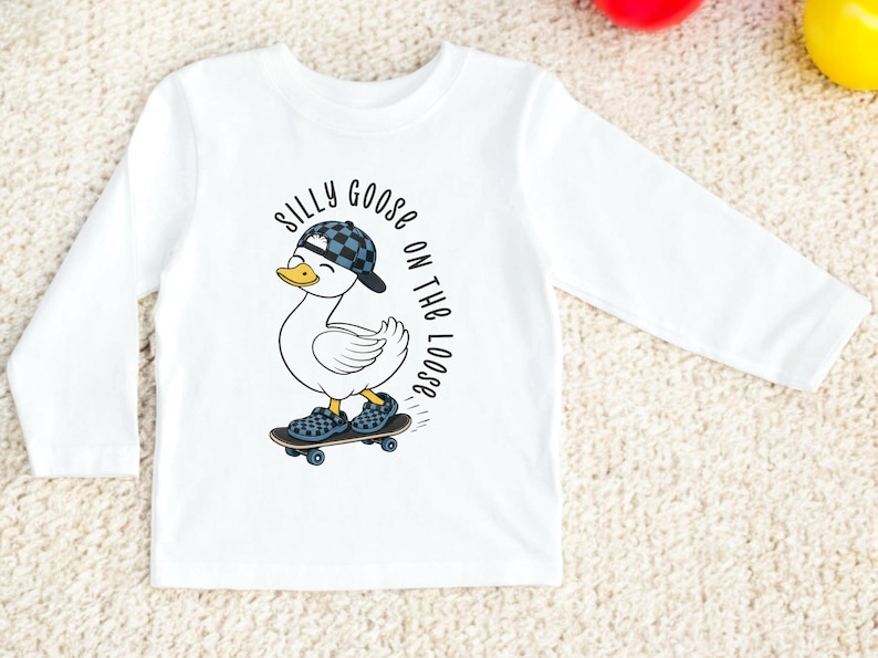 May include: White long-sleeved top with a cartoon duck wearing a cap and shoes, riding a skateboard. The text reads "Silly Goose on the Loose". The design is suitable for children's wear.