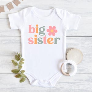 Puede incluir: Body de beb&eacute; blanco con la frase "big sister" en letras coloridas y un gr&aacute;fico de flores. El body est&aacute; sobre una superficie de madera blanca, con una ramita verde, un posavasos de madera y un mordedor.