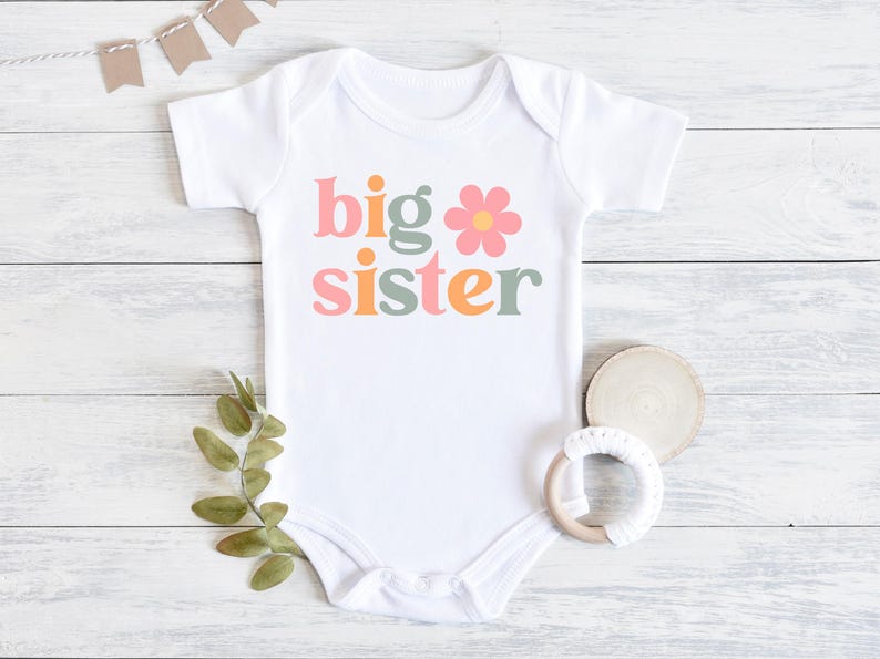 Puede incluir: Body de beb&eacute; blanco con la frase "big sister" en letras coloridas y un gr&aacute;fico de flores. El body est&aacute; sobre una superficie de madera blanca, con una ramita verde, un posavasos de madera y un mordedor.