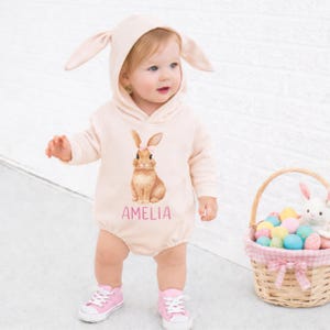 May include: A light pink baby romper with a bunny graphic and the name "AMELIA" in pink. The romper has a hood with bunny ears. A wicker basket with Easter eggs and a stuffed bunny is in the background.