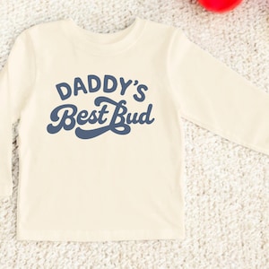 May include: Cream-colored long-sleeved shirt with the text "DADDY'S Best Bud" in navy blue script. The shirt is made of a soft material and is designed for children. The shirt is laying on a textured surface.