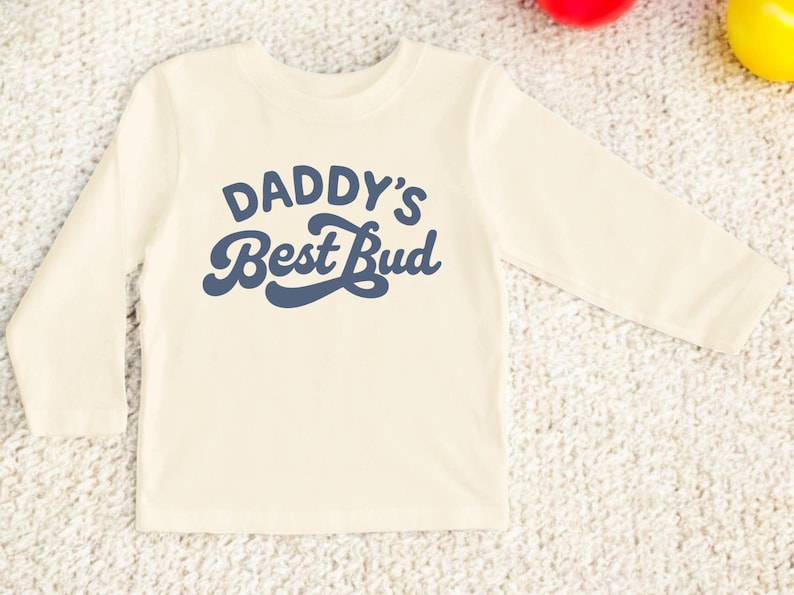 May include: Cream-colored long-sleeved shirt with the text "DADDY'S Best Bud" in navy blue script. The shirt is made of a soft material and is designed for children. The shirt is laying on a textured surface.