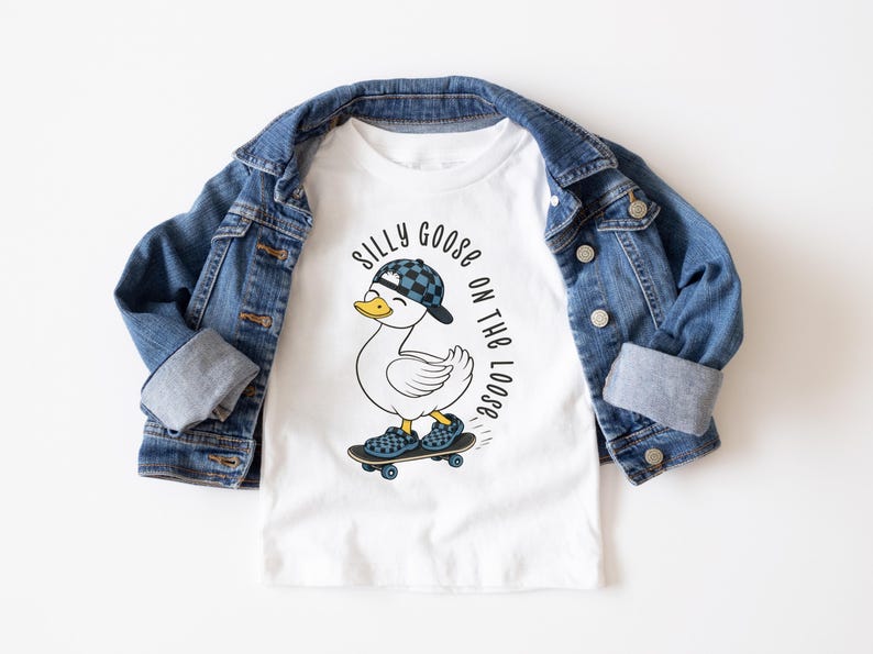 May include: White t-shirt featuring a cartoon goose on a skateboard, wearing a cap and shoes with a checkered pattern. The shirt is under a blue denim jacket. Text on the shirt says "Silly Goose on the Loose."
