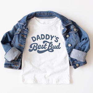 May include: A white t-shirt with the text "DADDY'S Best Bud" in a blue script font. The shirt is paired with a blue denim jacket. The jacket is buttoned and has rolled-up sleeves. The outfit is laid flat on a white surface.