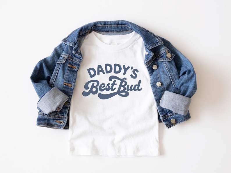 May include: A white t-shirt with the text "DADDY'S Best Bud" in a blue script font. The shirt is paired with a blue denim jacket. The jacket is buttoned and has rolled-up sleeves. The outfit is laid flat on a white surface.