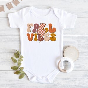 May include: White baby onesie with a retro-style graphic that says "Fall Vibes" in orange, brown, and pink letters with a checkered lightning bolt in the middle.