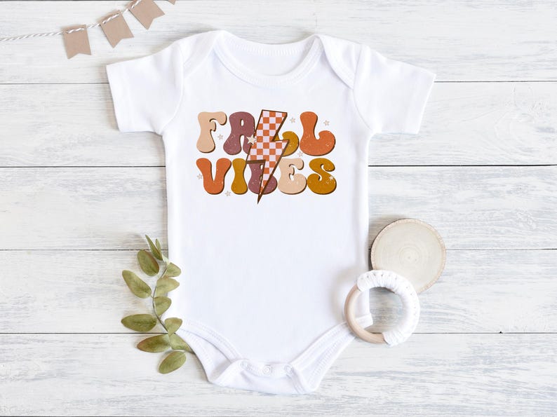 May include: White baby onesie with a retro-style graphic that says "Fall Vibes" in orange, brown, and pink letters with a checkered lightning bolt in the middle.