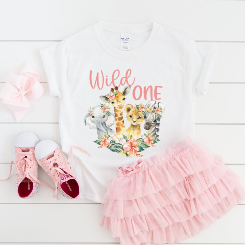 May include: White t-shirt with a pink "Wild One" graphic featuring a giraffe, lion, elephant, and zebra with floral accents. The shirt is paired with a pink tulle skirt.