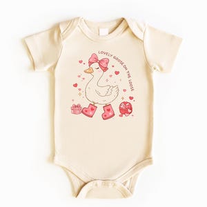 May include: Cream-colored baby bodysuit with a cartoon goose wearing a pink bow, boots, and surrounded by hearts. The text "LOVELY GOOSE ON THE LOOSE" is printed in a circular pattern. Includes gift boxes and heart-shaped candies.