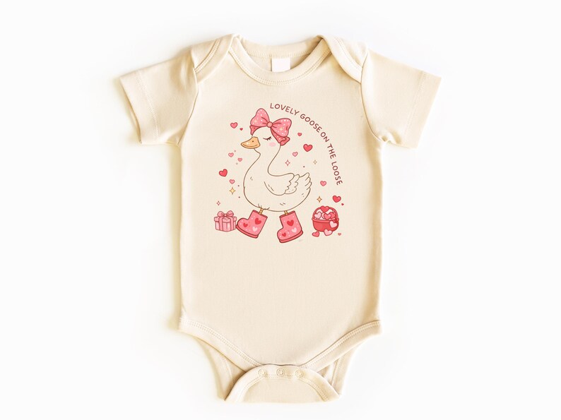 May include: Cream-colored baby bodysuit with a cartoon goose wearing a pink bow, boots, and surrounded by hearts. The text "LOVELY GOOSE ON THE LOOSE" is printed in a circular pattern. Includes gift boxes and heart-shaped candies.