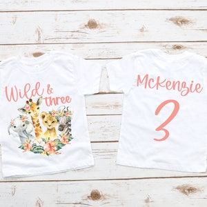 Puede incluir: Dos camisetas blancas con texto y gr&aacute;ficos rosas. La parte delantera de una camiseta dice "Wild & three" con una jirafa, un le&oacute;n, una cebra y un elefante. La parte trasera de la otra camiseta dice "Mckenzie 3".