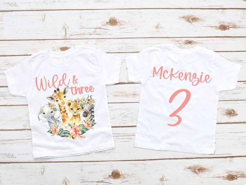 Puede incluir: Dos camisetas blancas con texto y gr&aacute;ficos rosas. La parte delantera de una camiseta dice "Wild & three" con una jirafa, un le&oacute;n, una cebra y un elefante. La parte trasera de la otra camiseta dice "Mckenzie 3".